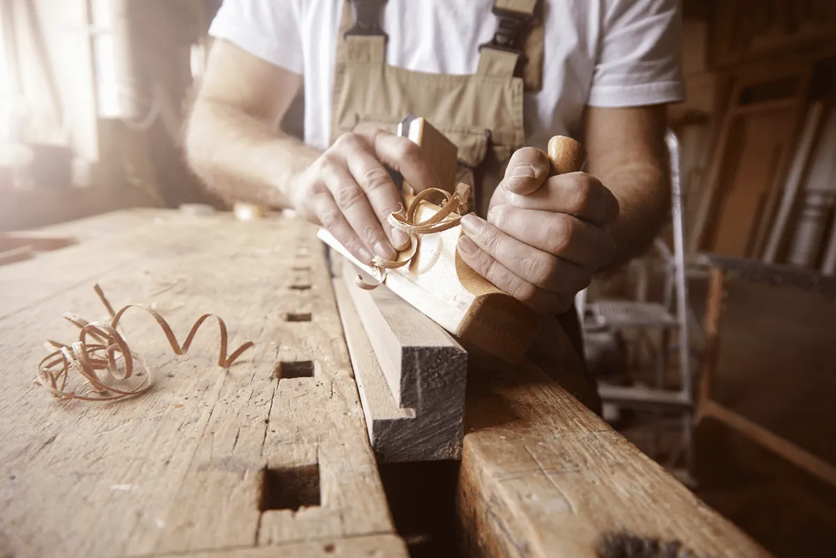 Close-up of hands guiding a hand plane along a timber board, with wood shavings on a workbench.