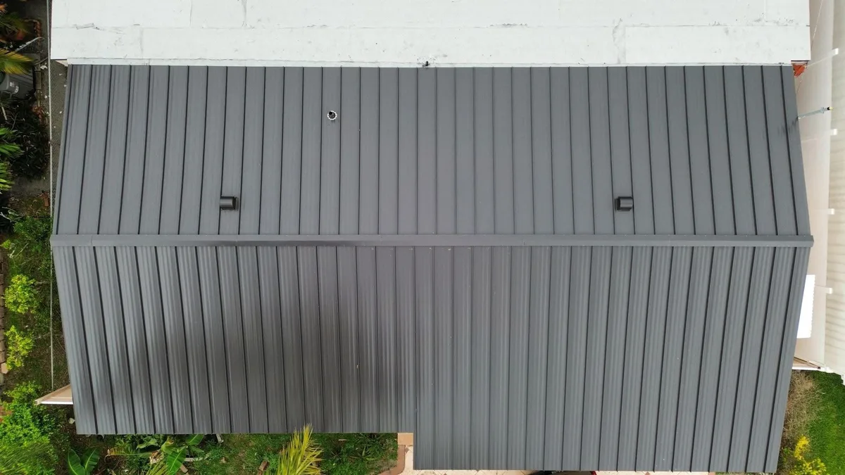 Overhead view of standing-seam metal roof panels, a ridge cap and roof penetrations beside a light-colored roof area.
